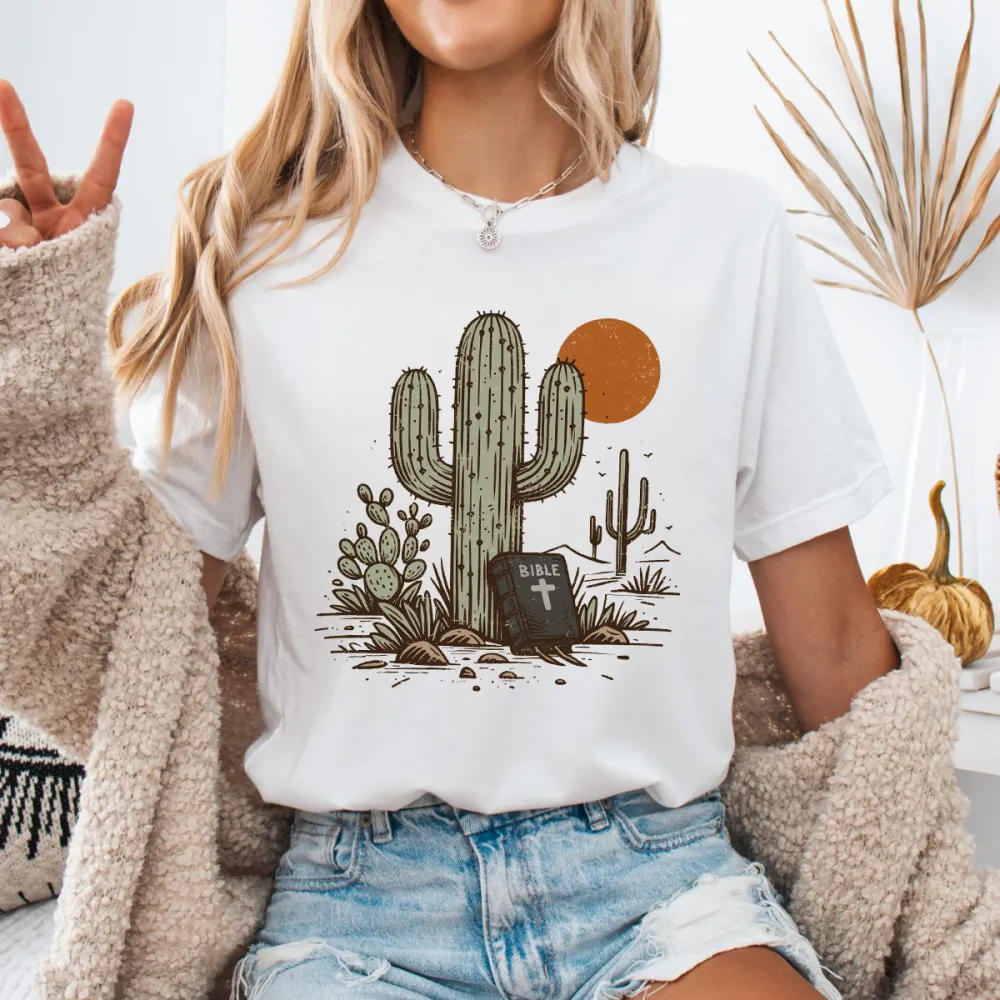 Cactus, With Small Cacti Next To It And A Tombstone With A Cross, With A Cactus Outline And An Orange Circular Sun Western Pattern In The Distance, Casual T-Shirt.