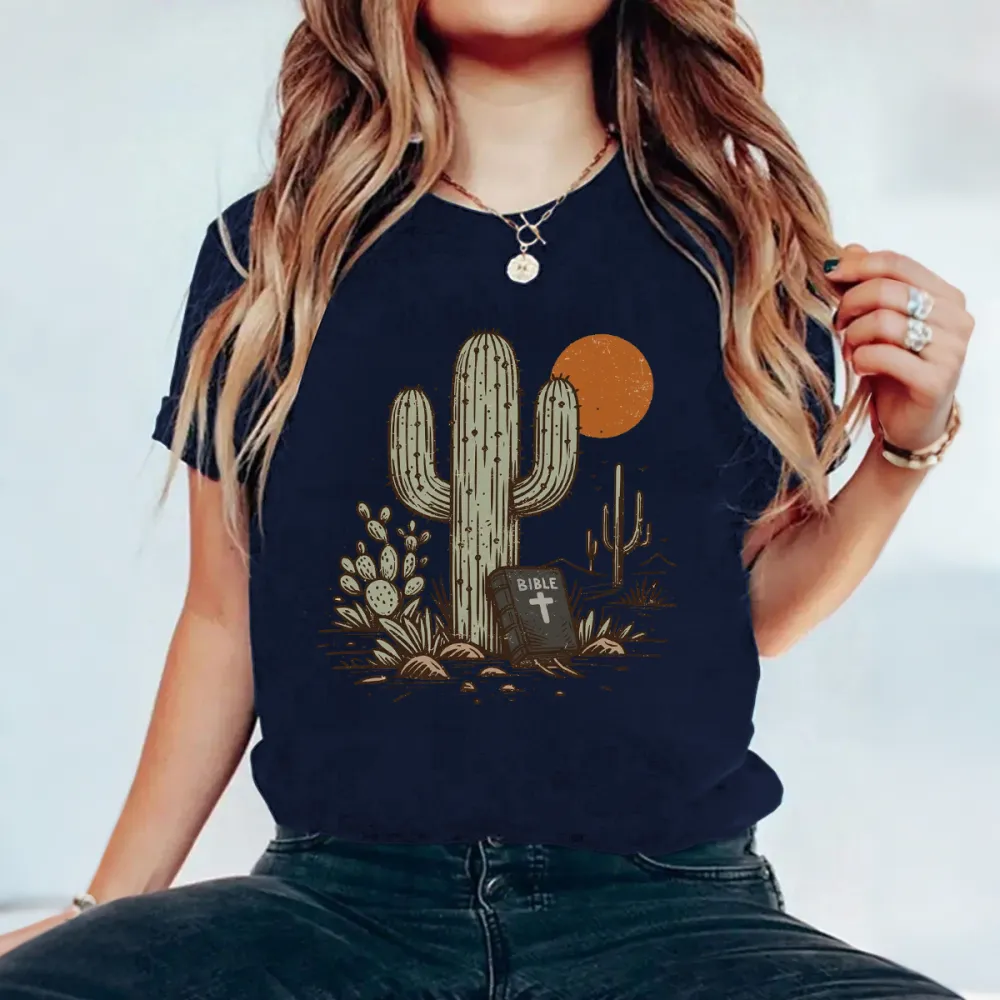 Cactus, With Small Cacti Next To It And A Tombstone With A Cross, With A Cactus Outline And An Orange Circular Sun Western Pattern In The Distance, Casual T-Shirt.